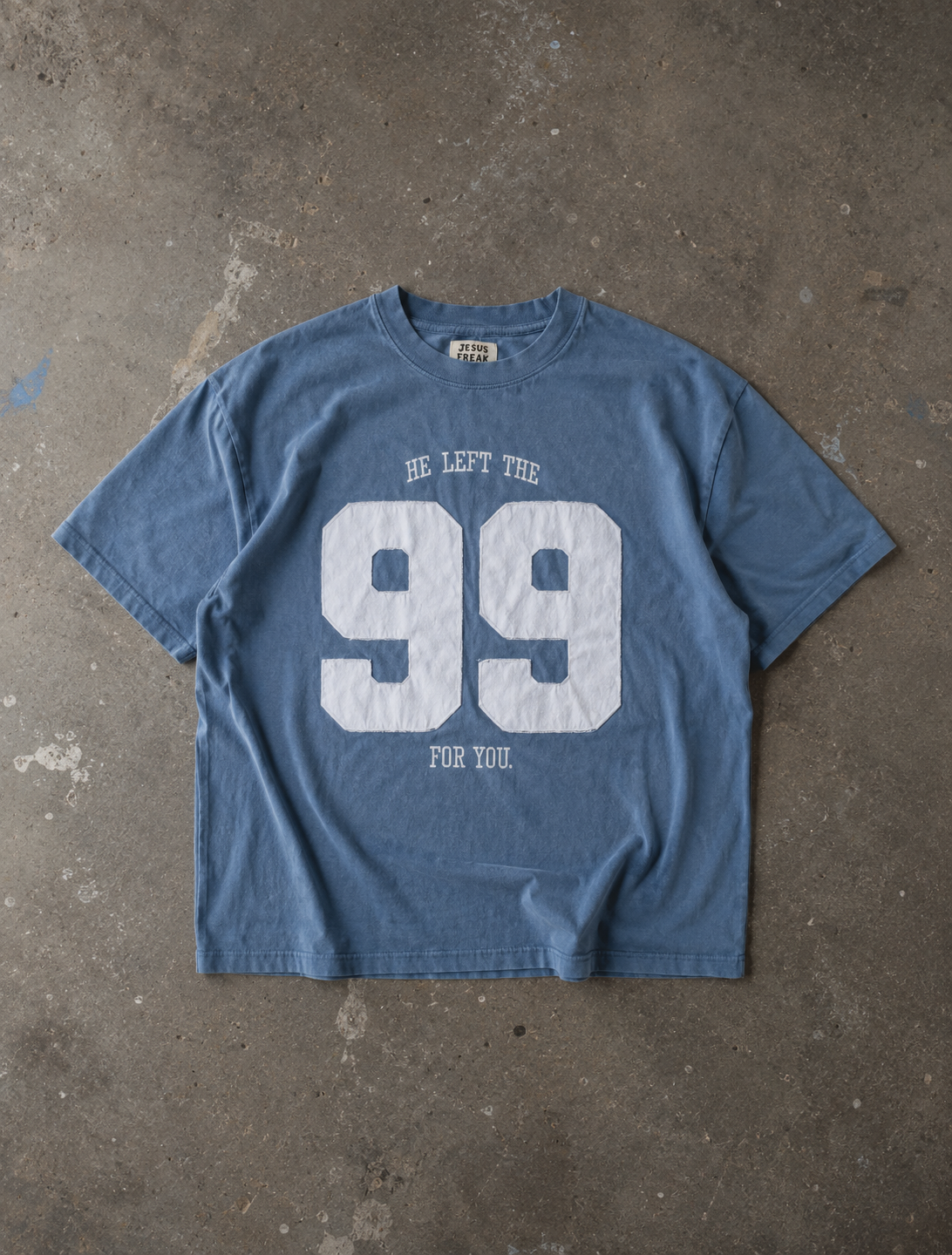 A faded blue graphic t-shirt with a distressed white print reading 'HE LEFT THE 99 FOR YOU' on the front, laid flat on a textured concrete surface.


