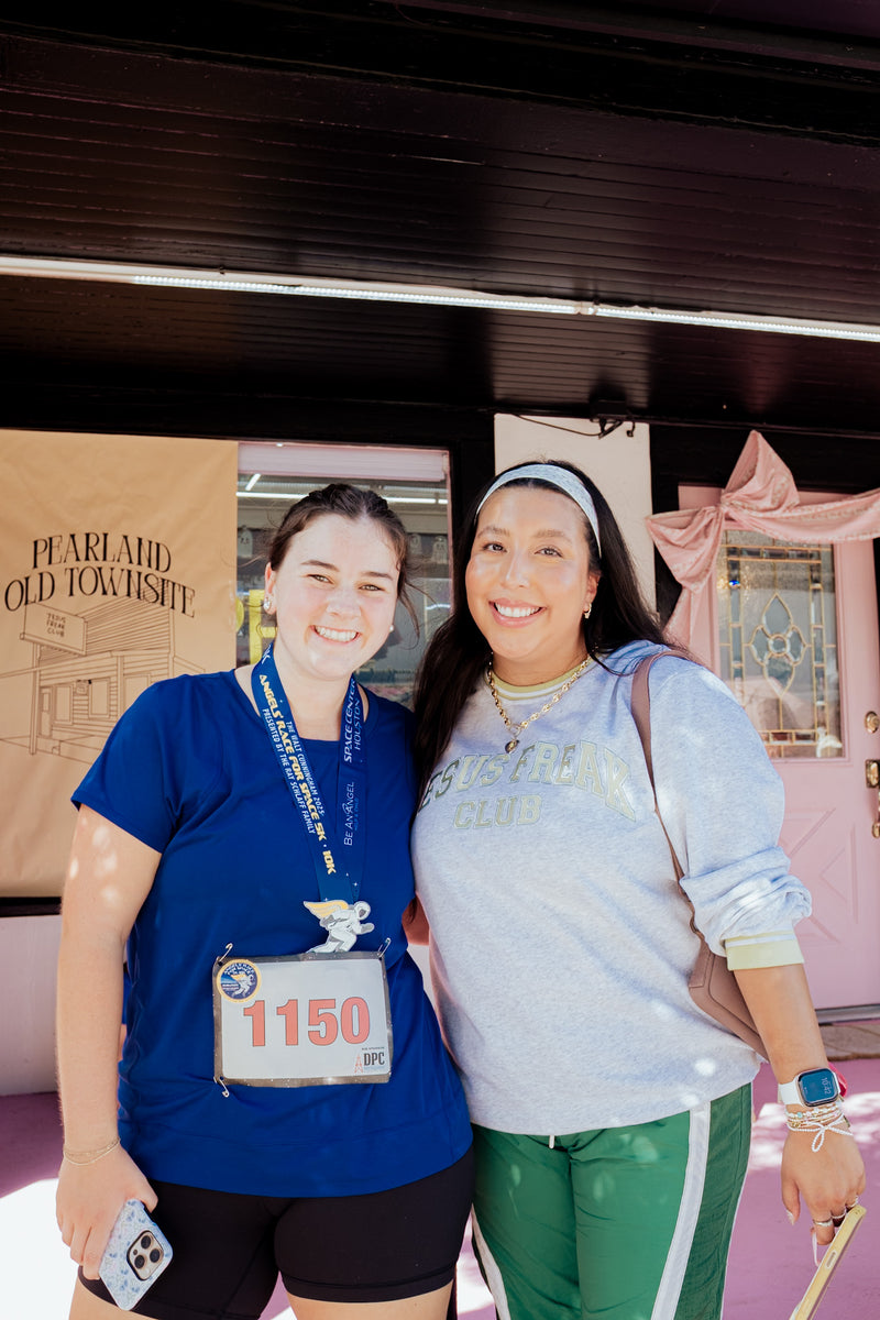 Two women smiling and posing together in front of a pink building entrance. The woman on the left wears a blue t-shirt, black shorts, and a race bib numbered 1150.


