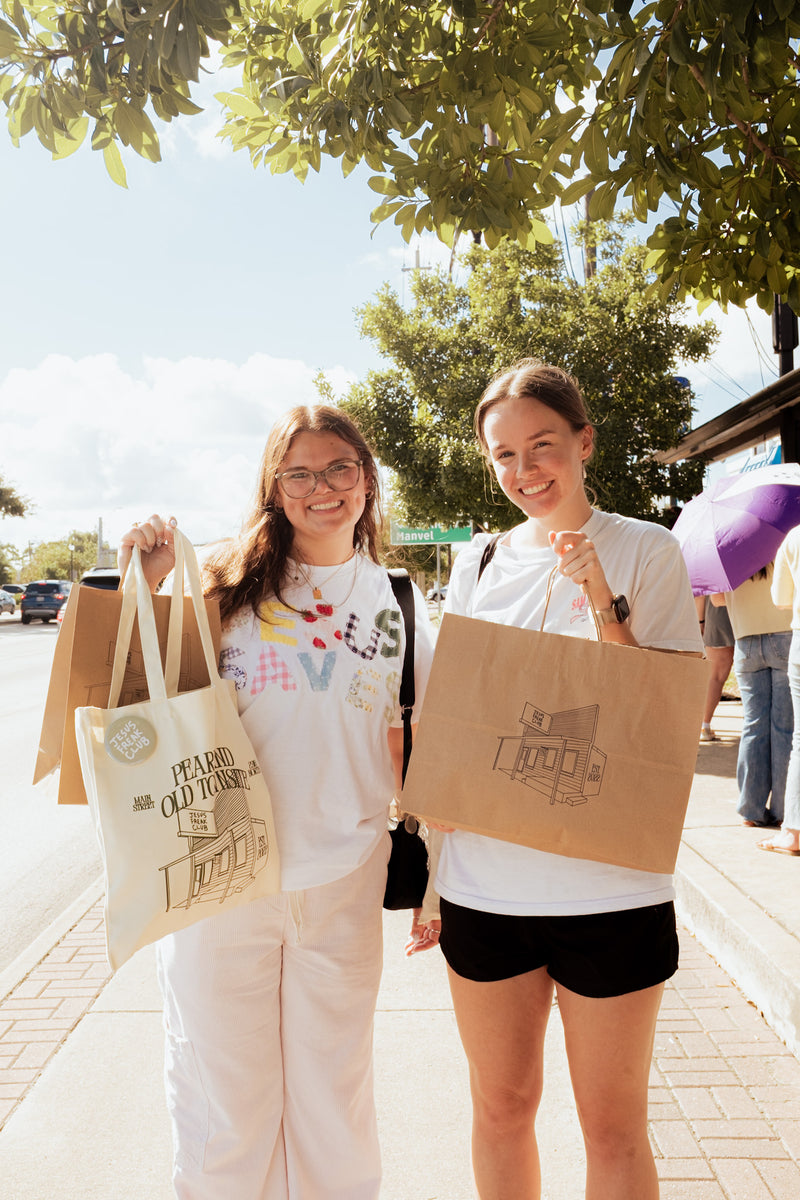 Two young women standing on a sunny sidewalk holding multiple paper shopping bags printed with the 'L.A.

