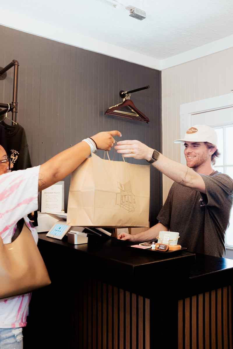 A person at a counter hands a brown paper shopping bag to a smiling delivery driver wearing a white cap and dark gray t-shirt in a modern retail setting.

